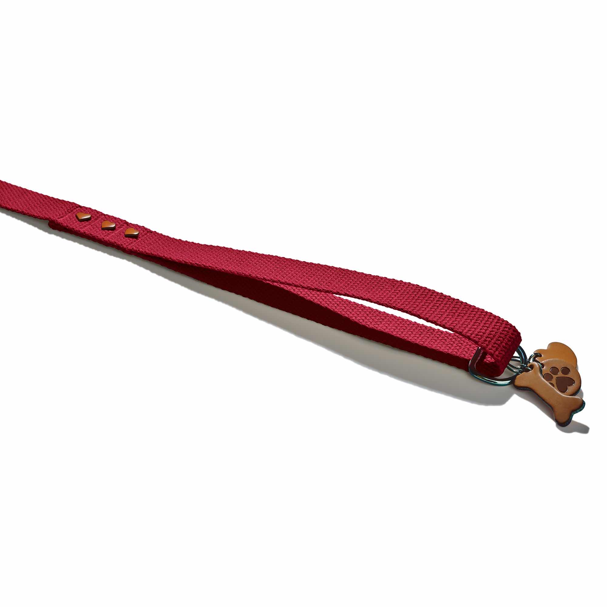 Gerader Leinengriff der Marke John Dog. Das Gurtband ist aus Baumwolle und hat eine kastanienbraune Farbe. Am Griff befinden sich drei herzförmige Nieten und ein Schlüsselring, auf dem ein Knochen, ein Herz und eine Hundepfote abgebildet sind.
